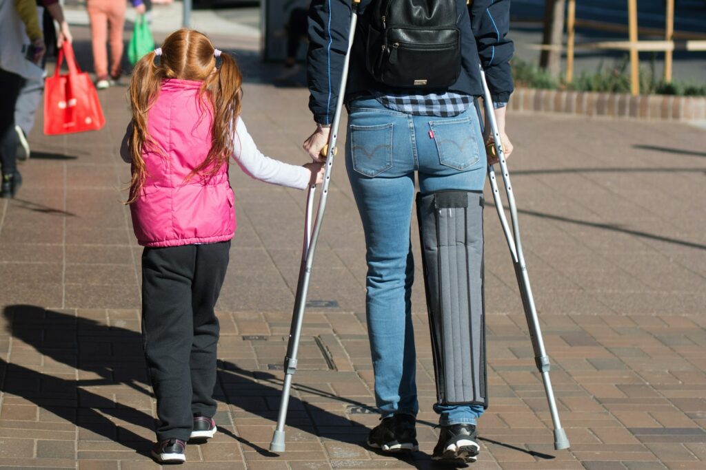 Woman with the crutches and knee brace on injured leg walking along the street with her daughter